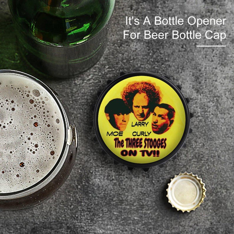 May include: A bottle opener with a black edge and a yellow center featuring Moe, Larry, and Curly from The Three Stooges. The text "The Three Stooges ON TV!!" is visible. The image also shows a beer bottle, a glass of beer, and a bottle cap.