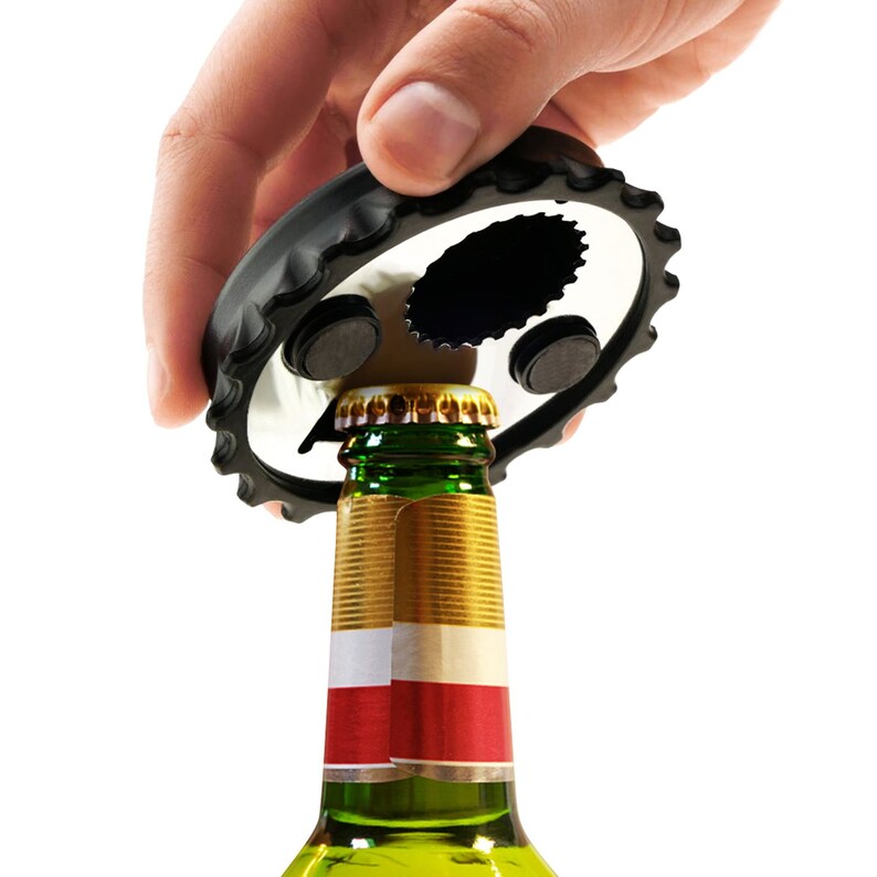 May include: A black bottle opener with a gear-like edge is positioned over a green glass bottle. The bottle has a gold cap and a red and white label. The opener is held by a hand.