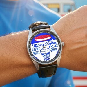 K&ouml;nnte beinhalten: Edelstahl-Armbanduhr mit schwarzem Lederarmband. Das Zifferblatt zeigt einen blauen Hintergrund mit den Worten "Mister Softee" und einer Eiscreme-Sandwich-Grafik. Die Uhr hat eine silberne L&uuml;nette mit schwarzen Ziffern.