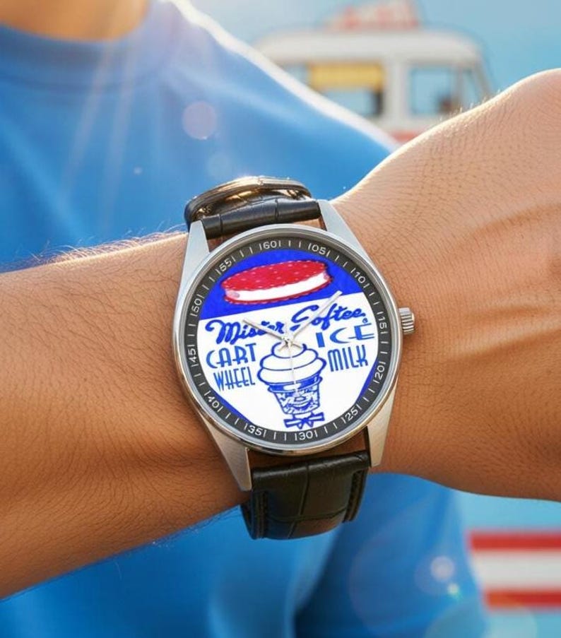 K&ouml;nnte beinhalten: Edelstahl-Armbanduhr mit schwarzem Lederarmband. Das Zifferblatt zeigt einen blauen Hintergrund mit den Worten "Mister Softee" und einer Eiscreme-Sandwich-Grafik. Die Uhr hat eine silberne L&uuml;nette mit schwarzen Ziffern.