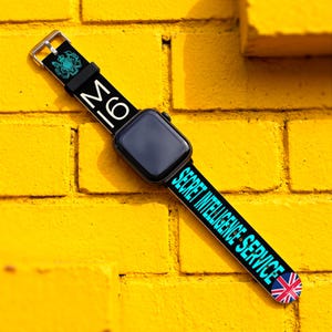 Peut inclure: Un bracelet de montre connectée noir avec le texte MI6 et Secret Intelligence Service en lettres blanches et cyan. Le bracelet comporte un emblème royal cyan près de la boucle argentée et un petit drapeau Union Jack à l'extrémité inférieure.
