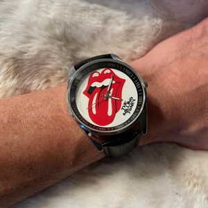 May include: A wristwatch with a black leather band and a silver-colored case. The watch face features the iconic red tongue and lips logo of The Rolling Stones against a white background. The watch has a black bezel with white minute markers.