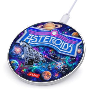 May include: A circular wireless charger with a silver rim, displaying the "Asteroids" logo and a space scene. The design includes a spaceship, planets, and the Atari logo. A white charging cable is attached.