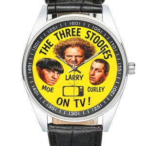 May include: A silver wristwatch with a black leather band. The watch face is yellow and features images of The Three Stooges, with the text "THE THREE STOOGES ON TV!" around the faces. The watch has a silver bezel.
