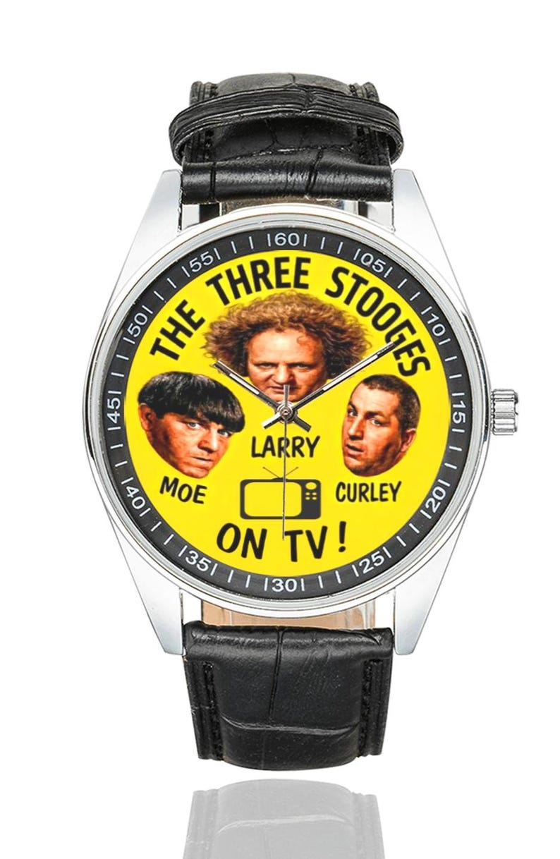 May include: A silver wristwatch with a black leather band. The watch face is yellow and features images of The Three Stooges, with the text "THE THREE STOOGES ON TV!" around the faces. The watch has a silver bezel.