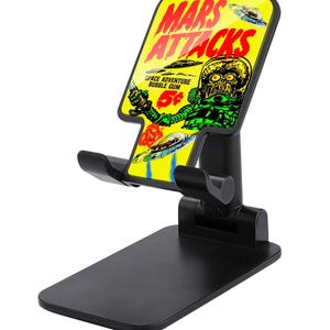 May include: Black adjustable phone stand with a yellow and red "Mars Attacks" graphic. The graphic features a green alien, flying saucers, and the text "Space Adventure Bubble Gum".