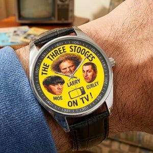 May include: A wristwatch with a silver case and a black leather band. The watch face is yellow and features images of The Three Stooges, with the text "THE THREE STOOGES ON TV!" around the images. An old television is in the background.