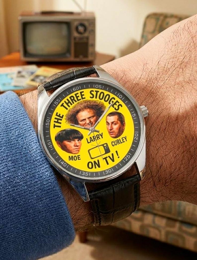 May include: A wristwatch with a silver case and a black leather band. The watch face is yellow and features images of The Three Stooges, with the text "THE THREE STOOGES ON TV!" around the images. An old television is in the background.
