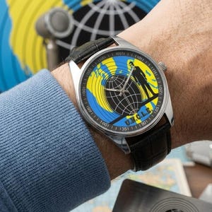 May include: A silver wristwatch with a black leather band. The watch face features a globe design in yellow and blue, with a silhouette of a person and the text "U.N.C.L.E."