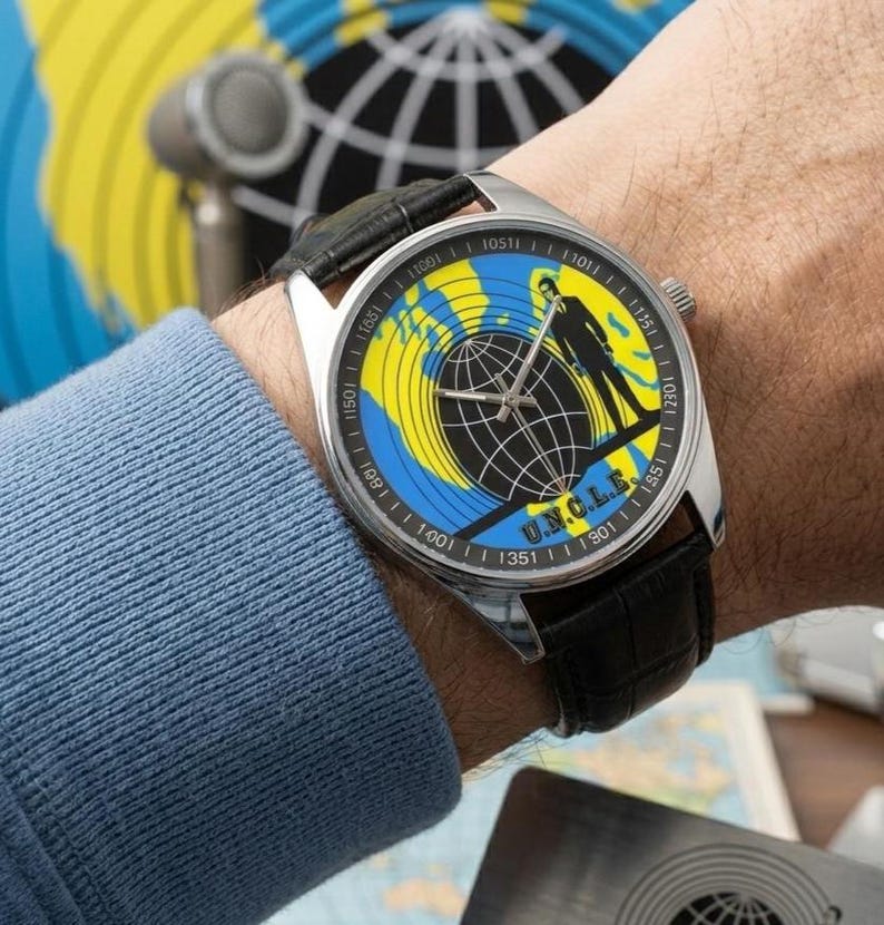 May include: A silver wristwatch with a black leather band. The watch face features a globe design in yellow and blue, with a silhouette of a person and the text "U.N.C.L.E."