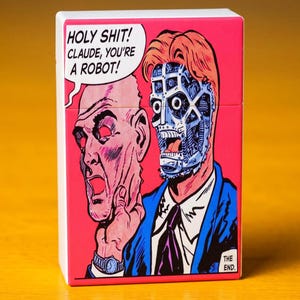 Può includere: Una scatola rettangolare con un'illustrazione in stile fumetto su sfondo rosa. Il disegno mostra una persona che rimuove una maschera umana per rivelare un volto robotico. Il testo nel fumetto recita: HOLY SHIT! CLAUDE, YOU'RE A ROBOT!