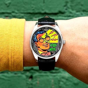 Op de afbeelding: Een polshorloge met een zwarte leren band en een zilverkleurige kast. De wijzerplaat toont een stripboekachtige illustratie van een man met een bril, met de woorden "RAY'S WATCH". Het horloge zit om een pols.
