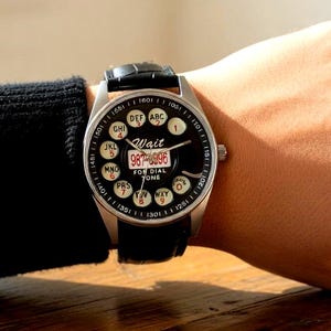 May include: A wristwatch with a black dial and a silver-colored case. The watch face features a rotary dial design with numbers and letters, and the text "Wait" and "FOR DIAL ZONE". The watch has a black leather band.