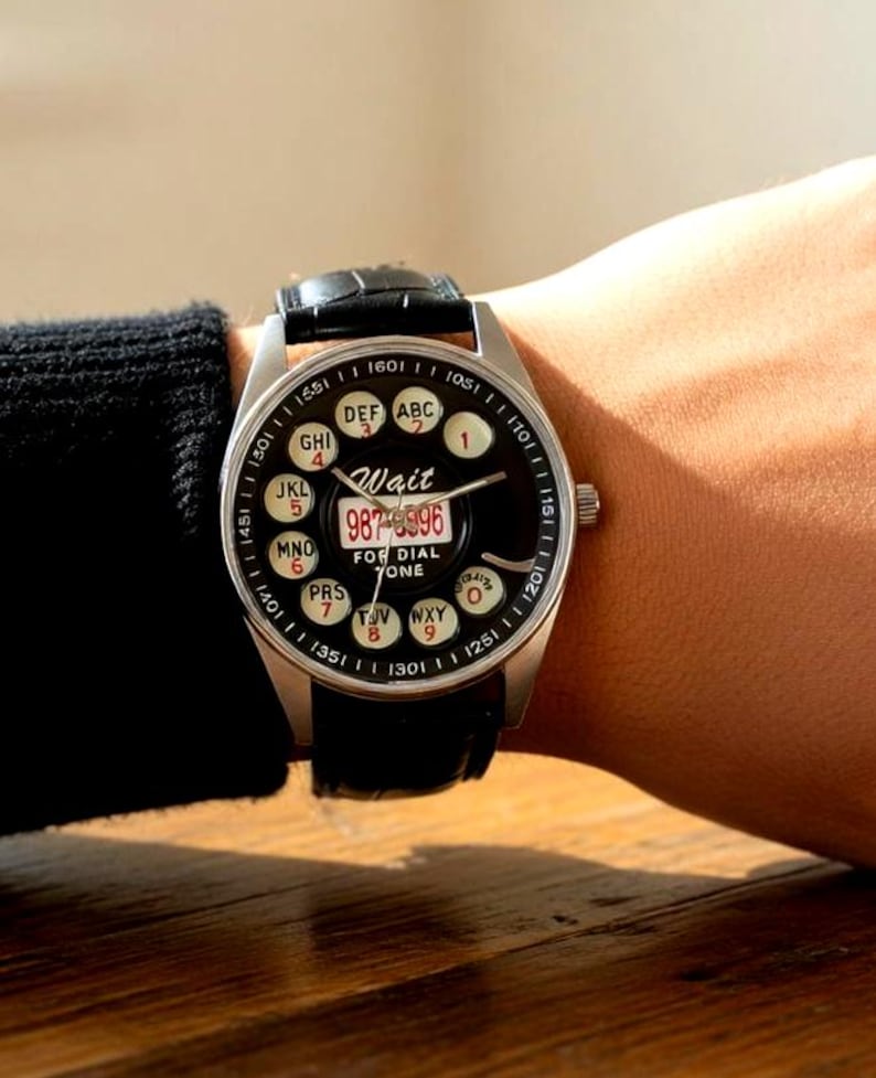 May include: A wristwatch with a black dial and a silver-colored case. The watch face features a rotary dial design with numbers and letters, and the text "Wait" and "FOR DIAL ZONE". The watch has a black leather band.