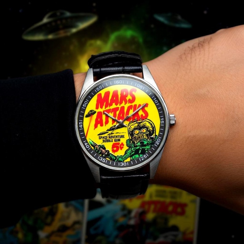 May include: A wristwatch with a silver case and a black leather band. The watch face features a yellow background with the words "MARS ATTACKS" in red and a green alien illustration. The watch also displays the text "SPACE ADVENTURE BUBBLE GUM 5&cent;".