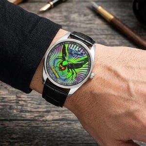 May include: A silver wristwatch with a black leather strap. The watch face displays a green and blue design with a stylized insect and the text "Work Hard, Play Harder".