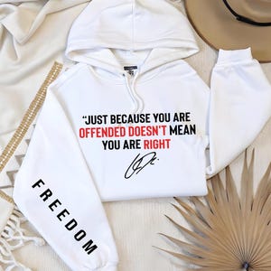 May include: White hoodie with the text "JUST BECAUSE YOU ARE OFFENDED DOESN'T MEAN YOU ARE RIGHT" in black and red. The sleeve reads "FREEDOM" in black. A signature is below the text. The hoodie has a drawstring hood.