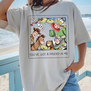 May include: Cream-colored t-shirt featuring a framed graphic of Toy Story characters, including Woody, Buzz Lightyear, and others. The text "YOU'VE GOT A FRIEND IN ME" is printed below the graphic. The shirt is a relaxed fit.