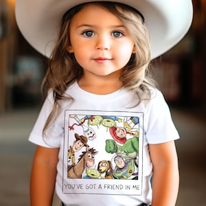 May include: A young child wearing a white cowboy hat and a white t-shirt. The t-shirt features a colorful graphic of characters from the movie "Toy Story" and the text "YOU'VE GOT A FRIEND IN ME".
