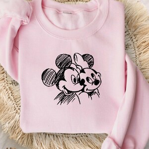 May include: A light pink sweatshirt featuring a black sketch of Mickey and Minnie Mouse. The sweatshirt has long sleeves and a crew neck. The design is centered on the front of the shirt. The sweatshirt is displayed on a woven, circular mat.