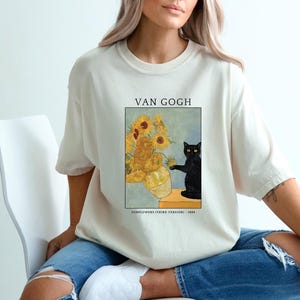 May include: Cream-colored t-shirt featuring a print of Van Gogh's "Sunflowers" with a black cat. The artwork includes a vase of sunflowers and a black cat. The text "VAN GOGH" and "SUNFLOWERS (THIRD VERSION) - 1888" are printed above and below the artwork.