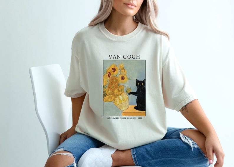 May include: Cream-colored t-shirt featuring a print of Van Gogh's "Sunflowers" with a black cat. The artwork includes a vase of sunflowers and a black cat. The text "VAN GOGH" and "SUNFLOWERS (THIRD VERSION) - 1888" are printed above and below the artwork.