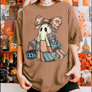 May include: A brown t-shirt featuring a whimsical ghost character in a patchwork outfit, a floral bow, and a pumpkin-shaped basket. The ghost has black eyes and is holding a blue pumpkin bucket. The shirt has a relaxed fit.