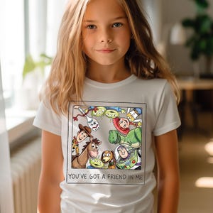 May include: White t-shirt featuring a colorful graphic of characters from the animated film "Toy Story." The design includes Woody, Buzz Lightyear, and other characters, framed by a black border. The text "YOU'VE GOT A FRIEND IN ME" is printed below the graphic.