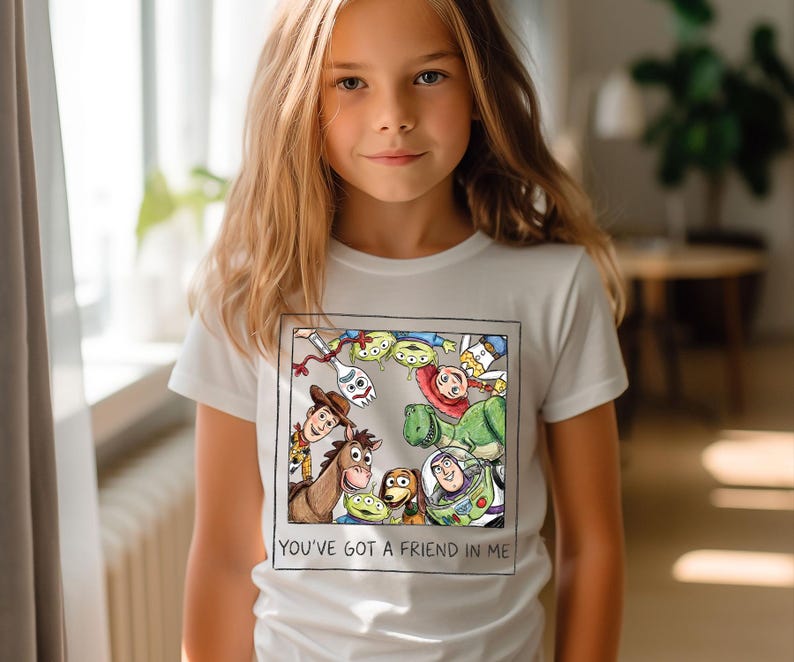 May include: White t-shirt featuring a colorful graphic of characters from the animated film "Toy Story." The design includes Woody, Buzz Lightyear, and other characters, framed by a black border. The text "YOU'VE GOT A FRIEND IN ME" is printed below the graphic.