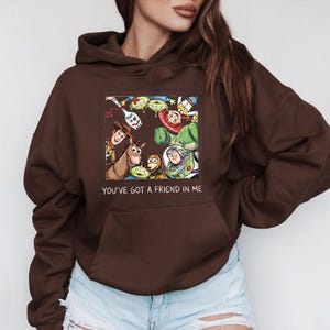 May include: A brown hooded sweatshirt featuring a colorful graphic of characters from the animated film "Toy Story." The graphic includes Woody, Buzz Lightyear, and other characters. The text "YOU'VE GOT A FRIEND IN ME" is below the graphic.