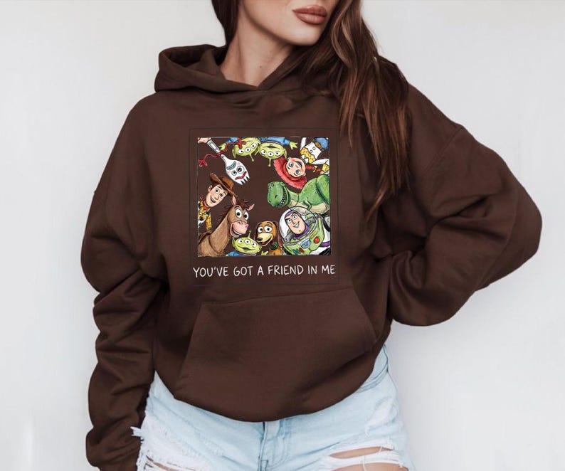 May include: A brown hooded sweatshirt featuring a colorful graphic of characters from the animated film "Toy Story." The graphic includes Woody, Buzz Lightyear, and other characters. The text "YOU'VE GOT A FRIEND IN ME" is below the graphic.