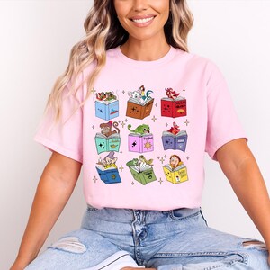 May include: A light pink t-shirt featuring a whimsical design of nine open books, each with a different cartoon character on top. The characters include a frog, a dragon, and a dwarf. The shirt is made of a soft material.