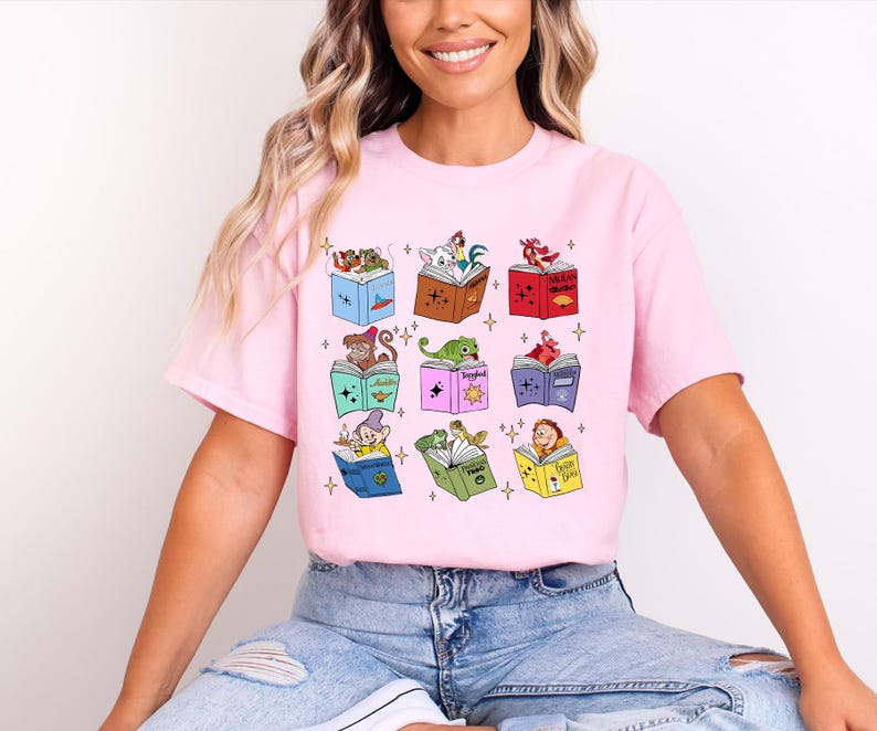 May include: A light pink t-shirt featuring a whimsical design of nine open books, each with a different cartoon character on top. The characters include a frog, a dragon, and a dwarf. The shirt is made of a soft material.