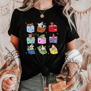 May include: Black t-shirt featuring a colorful graphic of nine open books, each with a different cartoon character reading inside. The books are in various colors, and the characters include a dog, a lizard, and Snow White.