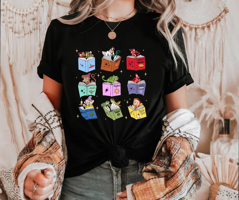May include: Black t-shirt featuring a colorful graphic of nine open books, each with a different cartoon character reading inside. The books are in various colors, and the characters include a dog, a lizard, and Snow White.