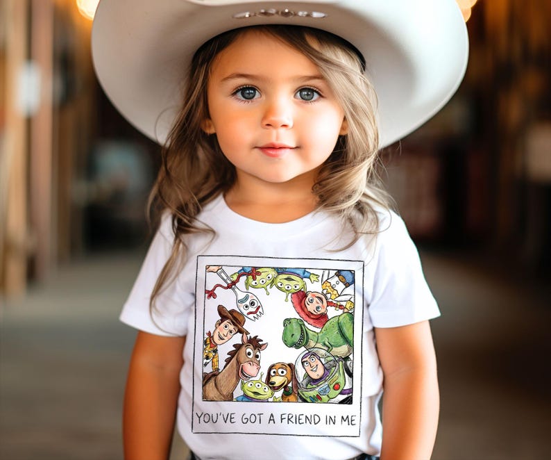 May include: A young child wearing a white cowboy hat and a white t-shirt. The t-shirt features a colorful graphic of characters from the movie "Toy Story" and the text "YOU'VE GOT A FRIEND IN ME".