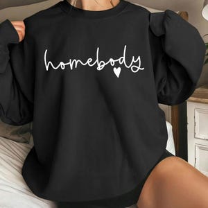 May include: Black sweatshirt with the word "homebody" in white script and a small white heart. The sweatshirt has long sleeves and a crew neck. The overall style is casual and comfortable.