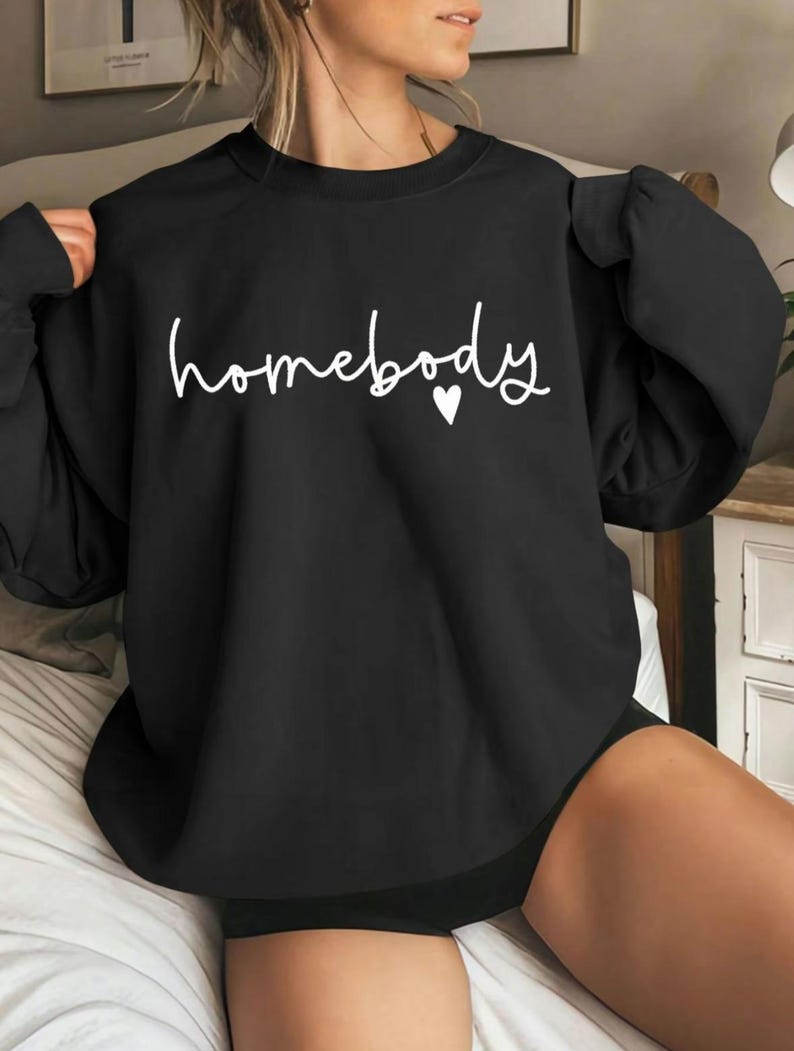 May include: Black sweatshirt with the word "homebody" in white script and a small white heart. The sweatshirt has long sleeves and a crew neck. The overall style is casual and comfortable.