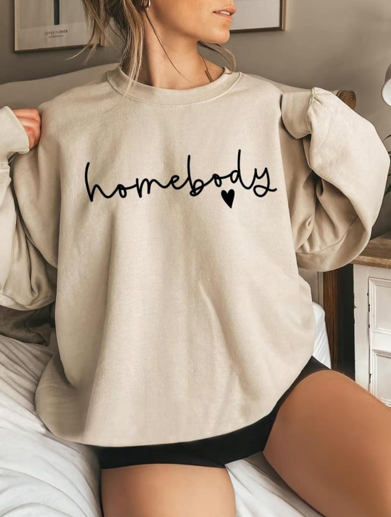 May include: A beige sweatshirt with the word "homebody" in black script and a small black heart. The long-sleeved crew neck top is made of a soft material.