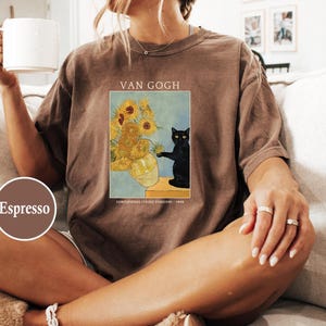 May include: Espresso-colored t-shirt featuring a print of Van Gogh's "Sunflowers" with a black cat. The shirt has short sleeves and a relaxed fit. The text "VAN GOGH" is above the image.