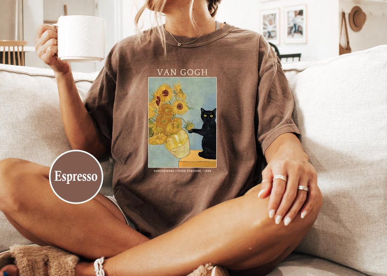 May include: Espresso-colored t-shirt featuring a print of Van Gogh's "Sunflowers" with a black cat. The shirt has short sleeves and a relaxed fit. The text "VAN GOGH" is above the image.