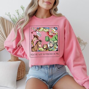 May include: Pink sweatshirt featuring a framed graphic of Toy Story characters. The graphic includes Woody, Buzz Lightyear, and the Little Green Men. The text "YOU'VE GOT A FRIEND IN ME" is below the graphic.