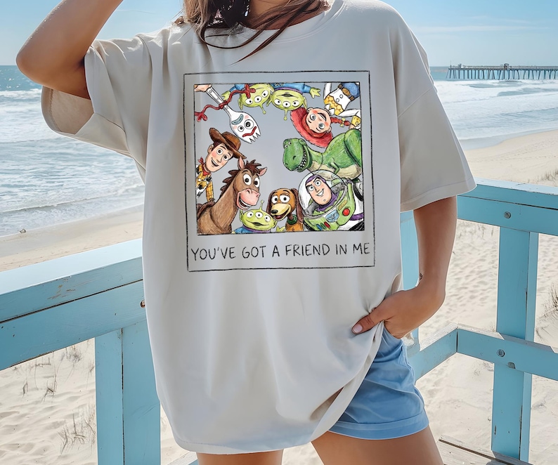 May include: Cream-colored t-shirt featuring a framed graphic of Toy Story characters, including Woody, Buzz Lightyear, and others. The text "YOU'VE GOT A FRIEND IN ME" is printed below the graphic. The shirt is a relaxed fit.