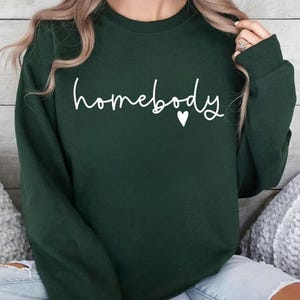 May include: A dark green sweatshirt with the word "homebody" in white script, a small white heart follows the word. The sweatshirt has a crew neck and long sleeves. The image shows the sweatshirt on a person.