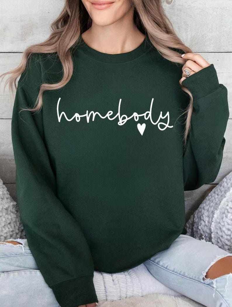 May include: A dark green sweatshirt with the word "homebody" in white script, a small white heart follows the word. The sweatshirt has a crew neck and long sleeves. The image shows the sweatshirt on a person.