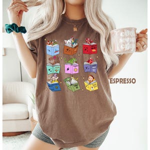 May include: Espresso-colored t-shirt featuring colorful illustrations of storybooks with various characters. The books are open, displaying images of a squirrel, frog, and other storybook characters. The word "Espresso" is printed in brown on the right side of the shirt.