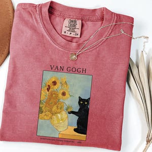 May include: A red-pink Comfort Colors t-shirt featuring a framed image of Van Gogh's sunflowers with a black cat. The shirt has a small tag at the neckline and a silver necklace. A brown hat and dried plant are also visible.