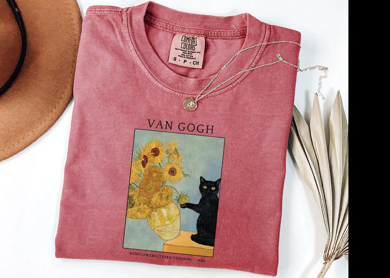 May include: A red-pink Comfort Colors t-shirt featuring a framed image of Van Gogh's sunflowers with a black cat. The shirt has a small tag at the neckline and a silver necklace. A brown hat and dried plant are also visible.