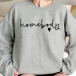 May include: A heather gray sweatshirt with the word "homebody" in black script, accompanied by a small black heart. The sweatshirt has long sleeves and a crew neck.