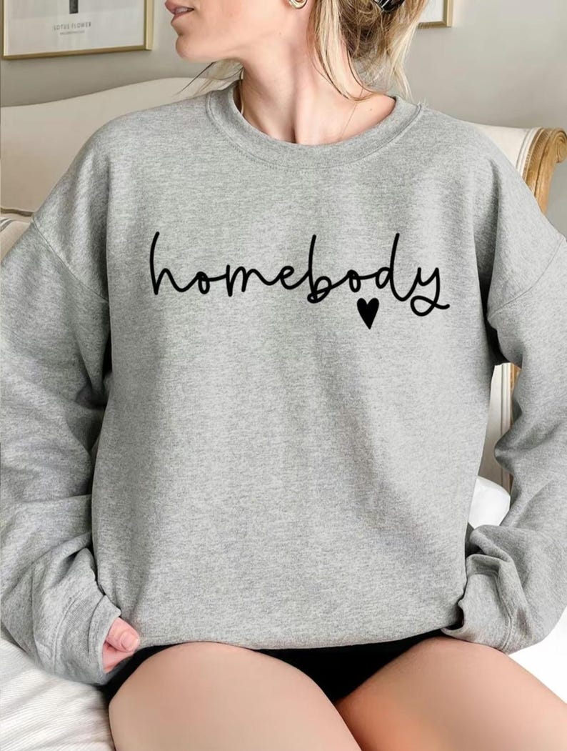 May include: A heather gray sweatshirt with the word "homebody" in black script, accompanied by a small black heart. The sweatshirt has long sleeves and a crew neck.
